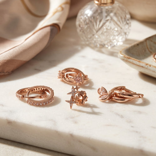 Four rose gold jewelry pieces, including rings and earrings, styled on a white marble tray with a glass perfume bottle and a beige scarf in the background.