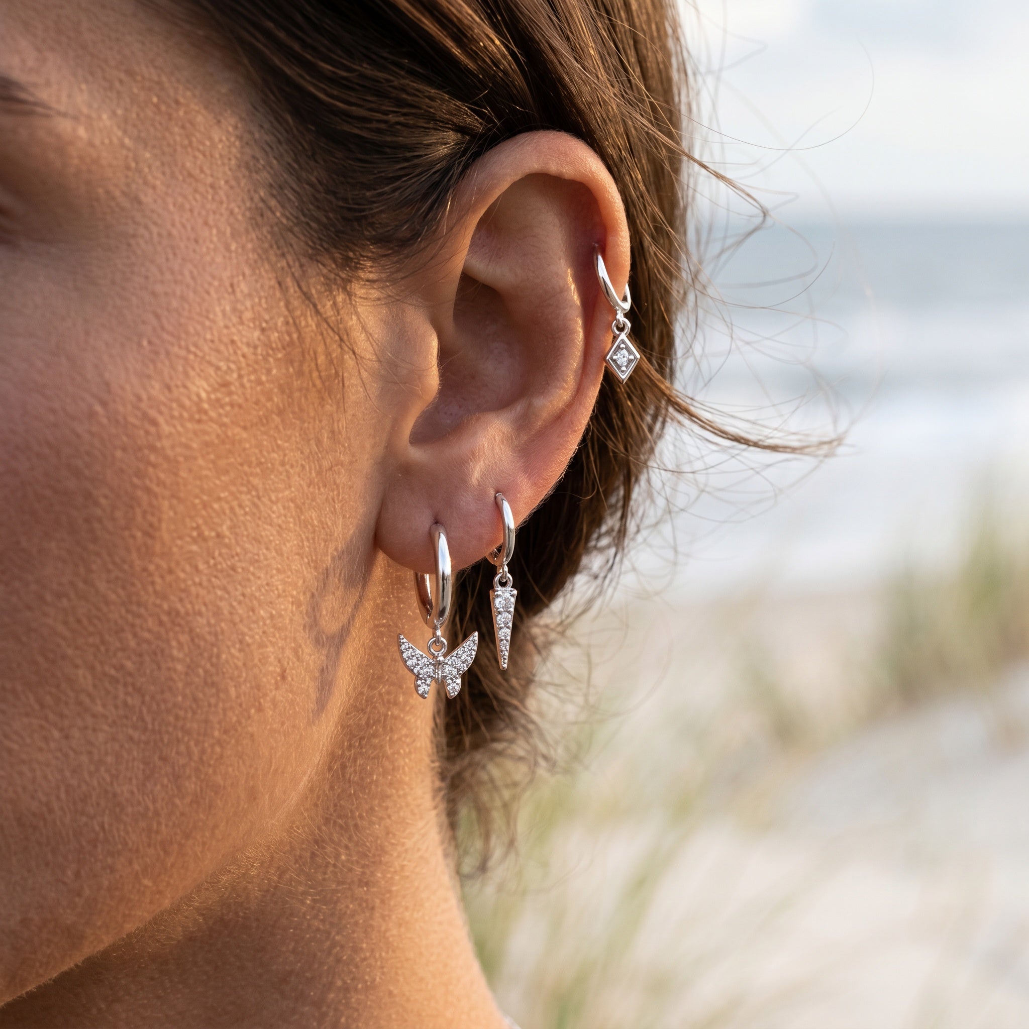 A close-up of a woman's ear wearing a set of silver hoop and dangle earrings with clear stones, set against a blurred beach background.