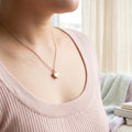 A close-up lifestyle shot of a person wearing a delicate rose gold heart pendant necklace on a fine chain, paired with a light pink ribbed knit top.