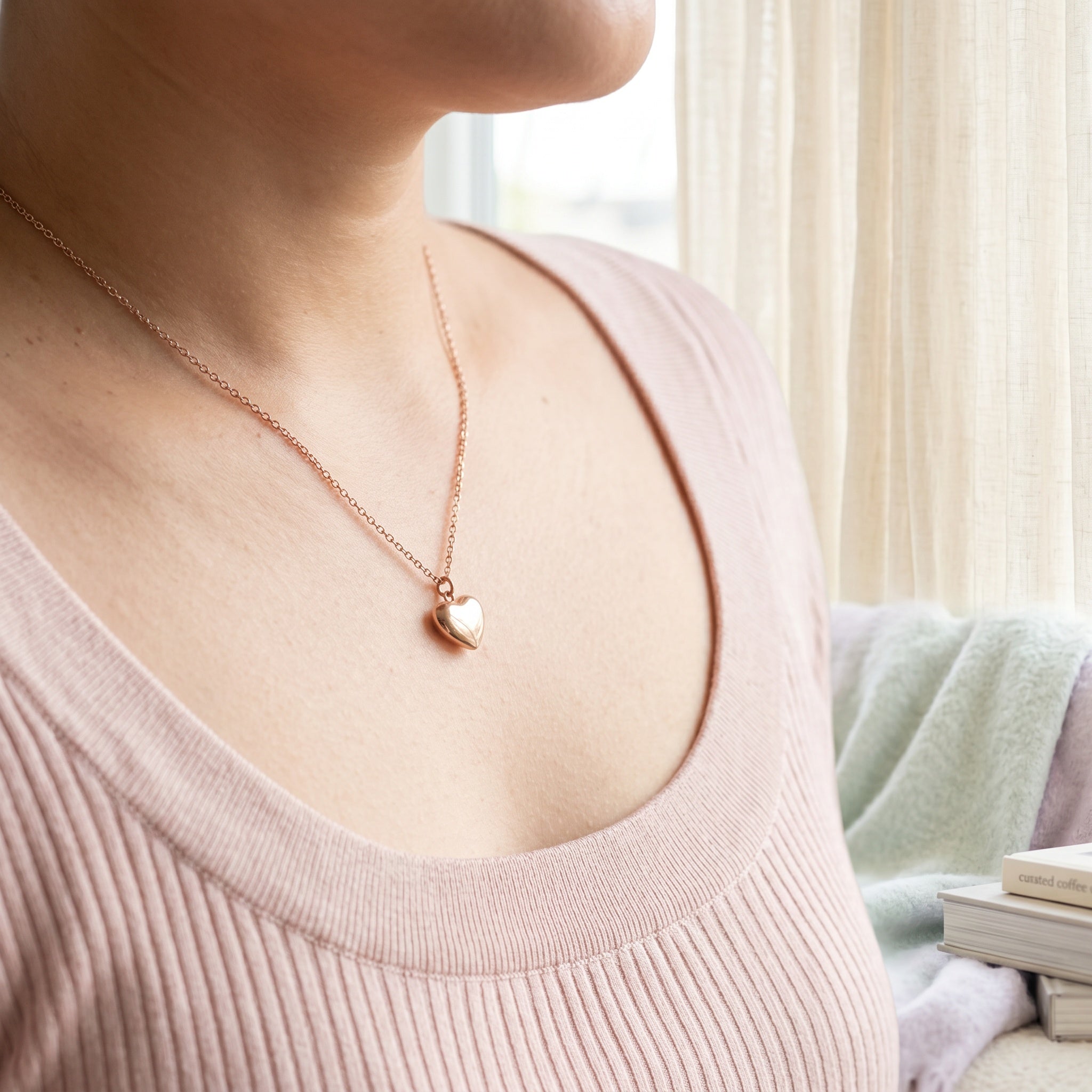 A close-up lifestyle shot of a person wearing a delicate rose gold heart pendant necklace on a fine chain, paired with a light pink ribbed knit top.